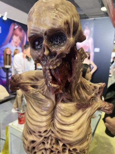 图片展示了一个极其逼真的恐怖风格怪物头部雕塑，位于某个展览现场。雕塑呈现出一种腐烂、扭曲的质感，皮肤呈现出病态的灰褐色，表面布满褶皱和孔洞。头部结构介于骷髅和某种未知生物之间，眼窝深陷且空洞，其中一只眼睛似乎有一个不规则的球体。嘴巴大张，露出不规整的牙齿和内部红色的组织。雕塑的颈部和肩部区域同样扭曲，肌肉和骨骼结构暴露在外，充满了令人不安的细节。背景中可以看到模糊的展览摊位和其他参观者，还有一个可口可乐罐放在雕塑旁边，提供了尺寸参考。整体氛围阴森恐怖，展现了高超的特效化妆或雕塑艺术技巧。