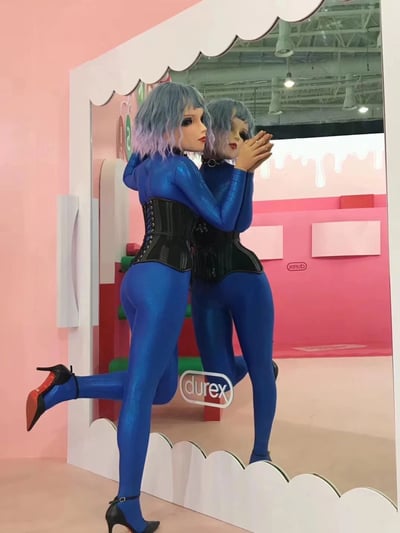 图中呈现杜蕾斯品牌线下展览的场景。一位模特身着亮蓝色紧身连体衣，外搭黑色束腰，蓝灰色渐变短发搭配精致妆容，正站在白色波浪边框的镜子前摆拍。她双手合十靠近脸颊，左腿向后抬起，姿态性感前卫，镜子清晰反射出她的背影。背景以粉色为主色调，墙面设有“yanjiuB”标识，地面可见“durex”品牌logo，整体场景色彩明快，充满时尚与品牌宣传的融合感。