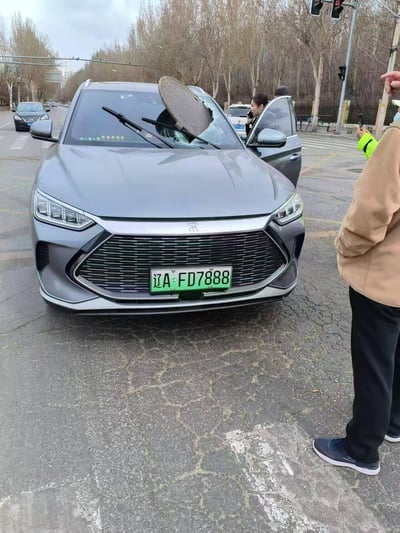 这是一张在城市路口拍摄的意外事故现场照片：一辆挂着辽A绿牌（辽A-FD7888）的灰色比亚迪宋新能源汽车，前挡风玻璃被一个圆形井盖直接砸穿，玻璃出现大面积蛛网式碎裂，井盖部分嵌入玻璃内。现场车门打开，一名女性在车旁打电话，还有穿着反光背心的工作人员及其他路人在旁围观，记录下这一罕见的井盖脱落砸中行驶车辆的惊险瞬间，场景让人感到意外和错愕。