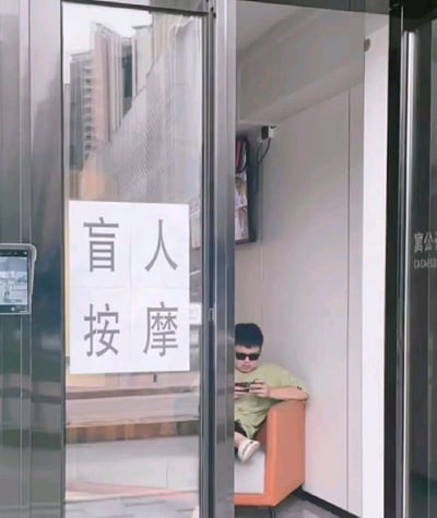 这张图片拍摄于一家写有“盲人按摩”招牌的小店门口，门上的纸条写着“盲人按摩”。门内沙发上坐着一位男生在低头玩手机，与外面招牌形成了明显的反差和幽默效果。重点在于“盲人按摩”的招牌容易让人产生歧义，本应为残障人士服务相关的场景却有普通人在休息，产生了无厘头的反转笑点。也暗含了广告语或现实场景与实际情况不符的常见网络玩梗方式。该场景属于生活中真实拍摄，因巧合和视觉误导引发网友二次传播。
