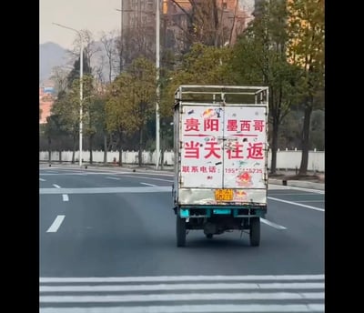 一张在城市道路上拍摄的照片，画面主体是一辆行驶中的三轮货车。车身后部有醒目的红色大字："贵阳 墨西哥 当天往返"，下方还有联系电话：195****5235。车身上贴有Red Bull的标志，车牌号为贵G·H27B。背景中可以看到树木、路灯和远处的建筑物及山脉。

这张图片的幽默之处在于将中国城市贵阳与北美洲国家墨西哥并列，声称可以当天往返，这在现实中是完全不可能的，形成了一种夸张的反差萌。