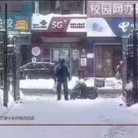 这是一张冬季雪天街头的实拍照片，画面中一名身着深色冬装的路人站立在街边商铺前，他身旁的雪堆因形状与位置巧合形成视觉错位，看起来如同有一个人倒在地上，制造出有趣的错觉效果。画面背景是带有“5G”“校园网办”字样的街边店铺，地面与车辆都覆盖着积雪，充满冬日氛围。