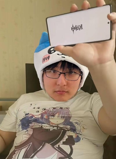 一个戴着白色蓝边猫耳帽子的人，身穿印有动漫少女图案的浅色T恤，正手持智能手机自拍。手机屏幕上显示着游戏《原神》的黑色logo。背景是带有花纹的浅色墙壁，整体氛围轻松日常，展现了游戏玩家的生活瞬间。