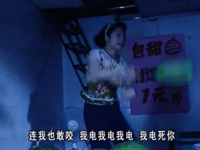 这是一张影视截图，画面呈现的是夜间卖西瓜的场景：一位中年女性穿着花色短袖，用发箍束起头发，她的表情激动愤怒，双手握着一个类似电蚊拍的器具不停挥舞，嘴中喊出“连我也敢咬 我电我电我电 我电死你”的话语，显然是因为被蚊虫叮咬后情绪激动，想要驱赶并“惩罚”蚊虫。她身后的紫色招牌上印有西瓜图案，还写着“包甜”和“1元/斤”的字样，表明这是一个西瓜售卖摊位，场景光线昏暗，充满了真实的市井生活气息，大姐的激动反应让这个日常场景显得格外搞笑生动。