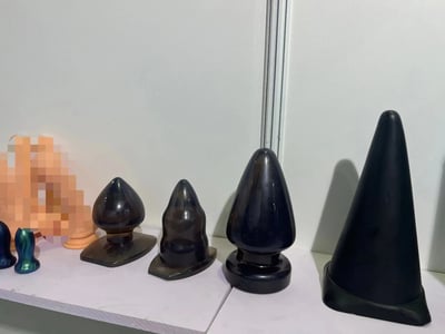 白色展示架上整齐排列着多个黑色锥形物体，表面光滑有光泽，形状略有差异，从左到右依次呈现从圆润到尖锐的变化。左侧有部分彩色物体被模糊处理。背景为纯白色墙壁，整体呈现简洁的展示场景。