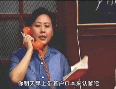 这是一张经过二次创作的影视梗图，画面主体是一位身着蓝色翻领衬衫的中年女性，她正将橙色老式有线电话贴在耳边通话，另一只手拿着一本翻开的红皮本子，表情严肃郑重，仿佛在传达一件极为正式的事情。画面下方的白色字幕写着“你明天早上带着户口本来认爹吧”，这句台词与人物严肃的神态形成强烈反差，制造出荒诞、搞笑的效果，打破了常规认亲场景的认知，常被网友在社交平台用来玩梗，以夸张的方式调侃认亲、发出邀约或制造戏剧化的玩笑氛围。