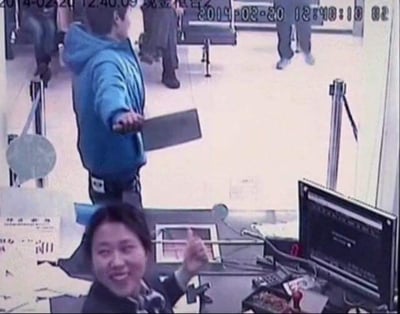 2014年2月20日的监控画面截图，显示一名身穿蓝色连帽衫的男子手持平板电脑指向柜台内，柜台后的女子正微笑着竖起大拇指回应。画面中有时间戳显示具体时间为12:40:09。