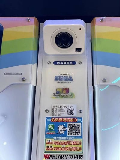图片展示了街机游戏《舞萌DX》(Wumeng DX)设备的玩家摄像头特写。中央为米色玩家摄像头，镜头呈黑色圆形，上方标注'玩家摄像头'。摄像头下方依次为：蓝色'SBGA'标志及日文'株式会社コナミ'(Konami Corporation)；彩色'舞萌 DX Chibi'游戏LOGO；白色设备标签，含'游戏游艺设备电子标识'字样、设备名称'舞萌DX(2021)'及二维码；底部红色边框的推广贴纸，内容为'免费获取玩家ID'，配有微信图标和二维码，标注'首次使用新玩家ID可以免费玩1首乐曲！'及'成人模式可以免费玩4首乐曲！'。设备左右侧板为彩虹条纹设计（红、黄、绿、蓝），整体呈现典型的街机设备视觉风格。