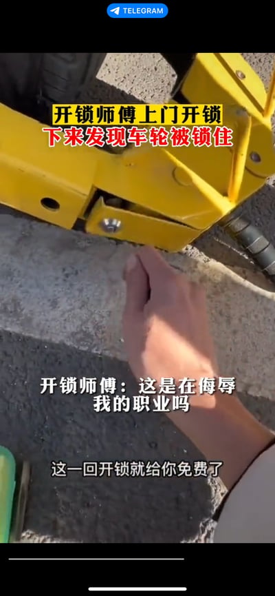 图片为近距离实拍场景，画面中心是一个黄色的车轮锁紧扣在地面，一只手正指向锁具。上方文字标注“开锁师傅上门开锁下来发现车轮被锁佳”（推测“佳”为“住”的笔误），下方配有开锁师傅的内心独白：“这是在侮辱我的职业吗 这一回开锁就给你免费了”。场景通过强烈的反差制造幽默效果——以开锁为职业的师傅，在提供服务时自己的交通工具反而被锁住，形成“专业人士遭遇同行（或恶作剧者）挑战”的讽刺画面，凸显生活中的荒诞与尴尬瞬间。
