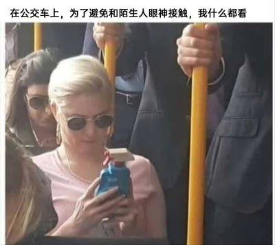 这是一张公交车内的实拍静态图，画面中一位身着粉色上衣、佩戴墨镜的女士，为了避免和周围的陌生人产生眼神接触，双手握着一个蓝色带喷头的清洁喷雾瓶，模仿握持手机的姿势低头专注地“看”着它，周围还有其他站立或坐着的乘客。图片顶部配有文字“在公交车上，为了避免和陌生人眼神接触，我什么都看”，精准调侃了社恐人群在公共场合，为了躲避和他人对视而做出的迷惑又真实的行为，充满搞笑与共鸣感。