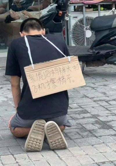 一名男子跪在户外砖石路面上，将头深深埋下，背上用白色绳子悬挂着一块手写硬纸板。纸板上用黑色字迹写着极具低俗羞辱性的内容：“我偷后妈裤头打飞机才擦精子。”，场景看起来是该男子因不当行为被处以羞辱式的公开惩罚，画面充满猎奇和低俗感。