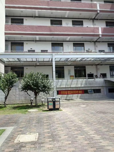 这是一张拍摄于国内某校园的实拍图，画面主体是一栋多层的校园建筑，底层墙面上悬挂着一条醒目的红色横幅，上面写着充满戏谑感的文字“劳资走啦!你们在这受苦吧!哈哈!”；在建筑二层的阳台处，有两名女生探身向外，似乎在互动。整体氛围轻松搞怪，是毕业生离校时用来调侃在校同学、抒发毕业解脱心情的玩梗内容。