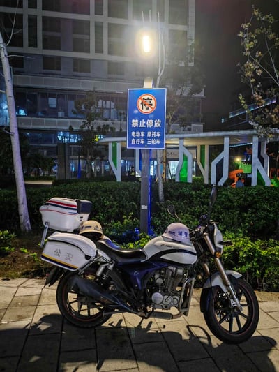 夜晚的户外场景中，一块蓝色的禁止停放标志牌明确标注了“禁止停放 电动车 摩托车 自行车 三轮车”，然而一辆带有“公安”标识的警用摩托车却直接停在标志牌前方的禁停区域内，形成了鲜明的规则反差。背景是现代化的商业建筑与绿化带，现场有灯光照明，能看到远处的工作人员和建筑内的灯光。