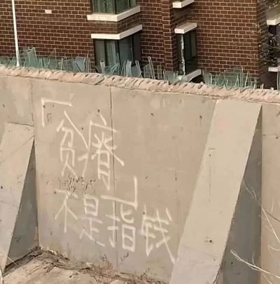 这是一张原始拍摄的静态单图，画面主体是一段顶部带有玻璃碴防护的水泥围墙，墙面上用白色涂料手写着“贫瘠不是指钱”的字样，字体风格较为随意。围墙后方是一栋红砖外墙的多层居民楼，楼体分布着多个带窗框的窗户，地面散落着干枯的杂草。图中的文字打破了大众对“贫瘠”一词通常关联物质金钱的固有认知，引导人们思考精神匮乏、认知局限等更深层次的“贫瘠”状态，带有一定的思辨性。