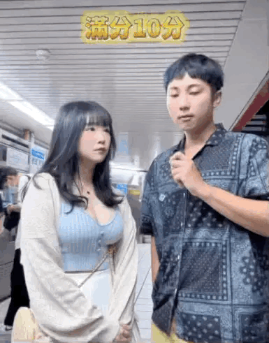 这是一张地铁内的街访视频截图，画面中有一男一女两位路人，上方有黄色文字标注“满分10分”，显然是在进行一场评分主题的街头采访。女生面露疑惑，看向身旁的男生，双手交握在身前；男生手持类似采访话筒的物品，神情专注，似乎正在提问或表达观点，场景位于地铁站这类公共交通空间，整体是生活化的街头互动场景。