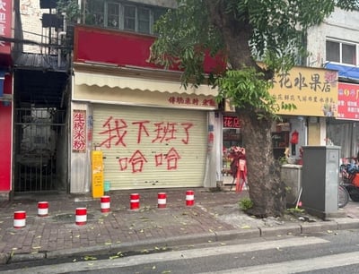 这张图片拍摄于一条城市街道的人行道旁。画面主体是一家已经拉下卷帘门、停止营业的米粉店。米白色的卷帘门上用醒目的红色喷漆随意地喷写着五个大字：“我下班了”，下方紧跟着“哈哈”二字，字体粗犷且充满喜感。店铺左侧的立柱上挂有“生榨米粉”的红色竖幅招牌，门头上也有“传统米粉、米饭”的字样。店铺右侧紧邻着一家名为“花水果店”的商铺，门口摆放着庆祝开业的花篮。人行道上排列着数个红白相间的圆柱形隔离墩，右侧有一棵枝繁叶茂的大树。整张图片通过店主随性幽默的“下班宣言”，展现了普通劳动者洒脱、接地气的一面，极易引发打工人的共鸣。