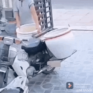 这是一段动态GIF，画面呈现了一处有趣的视觉错位场景：停在路边的摩托车，其后座结构与身旁地面上的白色水桶恰好重合，从特定角度看过去，仿佛摩托车“长”出了一个圆滚滚的肚子。旁边还有一位男子正手持另一个白色水桶，这个偶然形成的视觉错觉让日常场景变得十分滑稽搞笑。