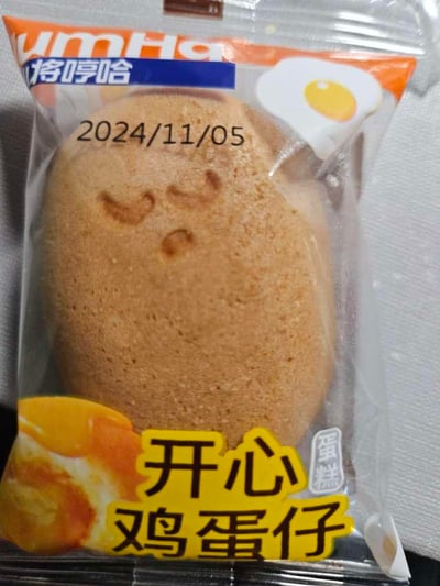 这是一张将哼哈品牌开心鸡蛋仔蛋糕的独立包装实拍图，包装为透明塑料材质，可清晰看到内部浅棕色的鸡蛋仔蛋糕，蛋糕表面压印出类似眯眼微笑的可爱表情。包装顶部印有品牌名“将哼哈”和生产日期“2024/11/05”，右下角标注“蛋糕”字样，底部用黄底黑字突出产品名“开心鸡蛋仔”，还搭配了流心蛋糕、煎蛋的图案，整体风格可爱，是一款适合作为早餐、下午茶的即食烘焙零食。
