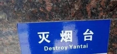 这是一张原始拍摄的横版图片，画面主体是一块带有蓝色边框的“灭烟台”标识牌，标识牌被固定在带有红黑纹理的深色石材墙面上。标识牌上方是白色的简体中文“灭烟台”，下方是对应的英文翻译“Destroy Yantai”，这是一处明显的错误翻译，将用于熄灭烟头的“灭烟”错误拆解，把“烟台”当成了地名，导致英文翻译出现“摧毁烟台”的荒谬含义，这种错误的翻译形成了滑稽的搞笑效果，让原本普通的公共标识变得充满趣味。