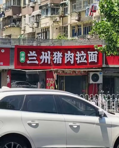 这是一张街头拍摄的实景照片，画面下半部分是一辆驶过的白色SUV，车辆后方是沿街的老旧居民楼，居民楼外墙上能看到晾晒的衣物、空调外机等生活设施。居民楼底层的餐馆使用红色门头招牌，招牌左侧印有绿色的清真标识，右侧是醒目的白色大字“兰州猪肉拉面”，内容存在明显的常识矛盾：清真餐饮有饮食禁忌严禁使用猪肉，而传统兰州拉面大多为清真品类，不会推出猪肉相关餐品，整体内容极具违和感。