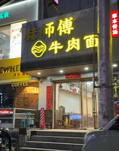 这是一张夜间拍摄的城市街边底商实拍照片，画面核心是“赫师傅牛肉面”的店铺门头：深色底的招牌上有黄色发光字体“赫师傅”“牛肉面”，二者中间是黄色的水波纹造型品牌logo。门头下方装有筒灯照明，店铺入口是玻璃推拉门，门口有几级台阶，门旁贴有红色对联类装饰，门上方贴着“生意興隆”的红色横批。店铺周边可见相邻的其他商铺，包括咖啡店、售卖草本骨汤的门店等，街边还停放着电动车、汽车，呈现出普通城市商业街的日常风貌。