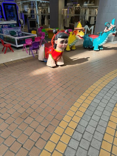 这是在商场外休闲区域拍摄的实景画面，地面铺设着棕、灰、黄三色拼接的地砖，阳光照射在地面形成清晰的阴影。画面中共有三台儿童摇摇车：最前方是哪吒造型的摇摇车，还原了哪吒标志性的双丸子头与红肚兜造型，表情呆萌；后方依次是戴着黄色帽子的黄色恐龙摇摇车、带有兔耳装饰的蓝色恐龙摇摇车。画面左侧设有一台标有“ideatank”字样的儿童钓鱼游乐池，池边摆放着红、粉、紫、黄等多种颜色的小型塑料椅，背景是橱窗内陈列着衣物的服装店，整体场景充满童趣，是商场为亲子家庭设置的儿童游玩区域。