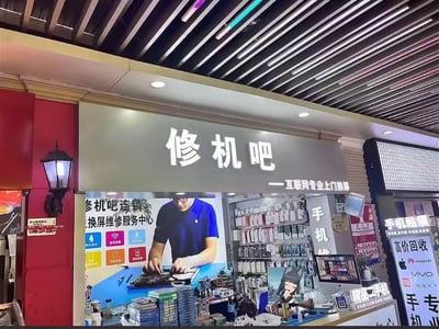 这是一家名为“修机吧”的手机维修店铺照片，拍摄于夜晚。门头招牌灯箱显眼，店内布置整齐，墙上陈列着各种手机配件，柜台前布满维修工具，还有品牌如HUAWEI、vivo、OPPO的标识。画面风格带有一点轻松幽默感，“修机吧”的店名容易让人联想到网络语“休息吧”，属于典型的双关梗，玩了一把谐音和休闲氛围。整体布置有浓郁的都市感和生活服务气息，适合用于吐槽手机坏了、休息片刻等场景。

- 名字易引发双关与谐音趣味：既能理解为“修理手机的地方”，又可被解读为“休息吧”这种轻松氛围。