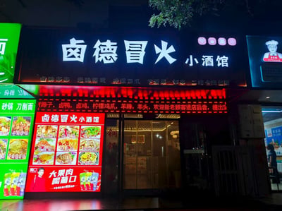 夜晚拍摄的「卤德冒火小酒馆」 exterior 照片，招牌以黑色为底，白色大字醒目，下方红色LED屏滚动显示菜品：卤味弟弟、绝味鸭货、柠檬鸡爪、下酒菜、烧烤等，二楼设卡座包间。左侧有绿色广告牌展示其他餐饮品类及菜品图片，右侧有厨师卡通形象logo。玻璃门入口可见店内灯光，整体呈现典型街边餐馆夜间亮灯场景。
