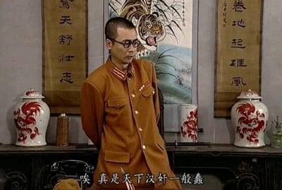 这是一张国产情景喜剧《地下交通站》的剧照截图，画面中是剧中经典汉奸角色贾贵，他身着土黄色的伪军制服，戴着眼镜，背着手站立在带有猛虎挂画和中式对联的厅堂背景前，脸上露出鄙夷、不屑的神情。画面底部配有字幕“唉 真是天下汉奸一般蠢”，贾贵本身是汉奸角色，却说出这样的台词，形成了强烈的荒诞讽刺效果，凸显了剧中对汉奸群体愚蠢可笑的刻画。