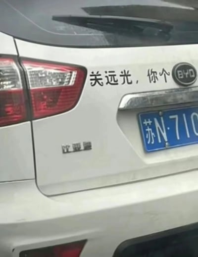 这是一张原始拍摄的静态照片，展示了一辆白色比亚迪汽车的尾部区域。车辆悬挂江苏宿迁（苏N）开头的蓝色民用汽车牌照，车尾右侧上方贴有未显示完整的黑色文字标语“关远光，你个”，意在提醒后方车辆不要滥用远光灯，属于车主自行张贴的个性化车贴；车尾带有比亚迪的英文品牌标识“BYD”和汉字“比亚迪”字样，车辆的红色尾灯、后挡风玻璃雨刷等部件清晰可见，车身表面有轻微的灰尘污渍。