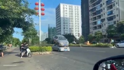 这是一张在城市街头拍摄的照片，两辆同款银色轿车因拍摄角度巧合，看起来像一辆叠在另一辆车顶，形成滑稽的视觉错位效果。画面左侧非机动车道上有骑电动车的路人，背景是居民楼和挂红灯笼的路灯，日常街景因这个错位变得十分有趣。
无谐音梗或双关，仅为拍摄角度造成的视觉错觉。