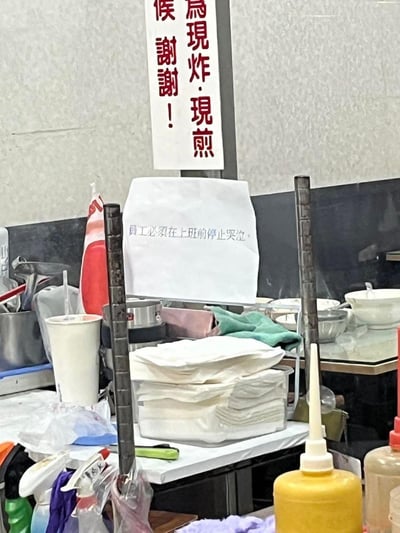 这是一张餐饮店铺操作区的实拍图，操作台上堆放着纸巾、饮品杯、餐具、清洁布等杂物，背景的操作区域也摆放着各类厨具。画面中有两块标语牌：上方是繁体文字的“为现炸·现煎 候 谢谢！”，是店铺告知顾客餐品为现制的提示；下方的白纸上用简体字写着“员工必须在上班前停止哭泣”，这句直白又心酸的标语，带着黑色幽默的意味，直观展现出餐饮从业者的工作压力与职场的无奈。图中没有双关、谐音梗等内容。