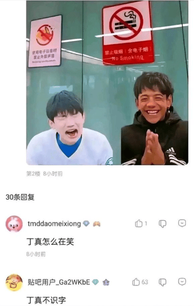 这是一张来自百度贴吧的社交媒体截图，截图主体是一张网友合成的图片：画面中有两位男性，左侧男子表情激动，大张着嘴，身体前倾；右侧是丁真，他面带灿烂笑容，双手合十放在身前。两人身后的玻璃墙面上贴着两个标识，左侧标识带有禁止电子设备外放的图案，下方标注文字“使用电子设备时禁止外放声音”；右侧是国际通用的禁止吸烟标识，下方标注“禁止吸烟（含电子烟）No Smoking”。截图下方是两条网友评论，第一条用户“tmddaomeixiong”提问“丁真怎么在笑”，第二条用户“贴吧用户_Ga2WKbE”回复“丁真不识字”，这条回复获得了63个点赞，该评论是玩梗调侃丁真不认识标识上的文字，从而制造出丁真因为看不懂标识而开心的搞笑效果。