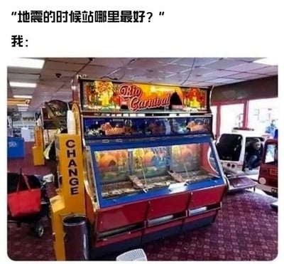 这是一张图文混排的静态搞笑梗图，上方配有文字提问“地震的时候站哪里最好？”，随后以“我：”引出下方的游戏厅场景图。图中展示的是一台名为“Rio Carnival”的大型推币游戏机，机器主体为红蓝色，外观装饰有色彩鲜艳的热带风格图案，旁边还设有标有“CHANGE”字样的黄色兑币装置，整体场景是典型的室内游戏厅环境。这张图通过幽默的方式回应了地震避险的问题，暗示地震时可以躲在这类结构坚固、重量较大的重型设备下方，用轻松搞笑的形式传递地震避险的常识，属于网络上流行的问答式梗图。