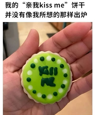 图片上方配有文字‘我的“亲我kiss me”饼干并没有像我所想的那样出炉’，下方是一只手握着一块绿色糖霜饼干的实拍图。饼干表面覆盖亮绿色糖霜，边缘有绿色和白色圆点装饰，中央试图用深绿色糖霜写出‘kiss me’字样，但字迹潦草模糊无法辨认，呈现明显烘焙失败效果，整体氛围搞笑又无奈。