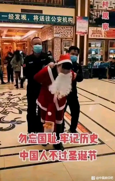 这张图片拍摄于装修精致的商场大厅，地面带有花纹地砖，背景可见店铺与往来行人。画面中两名身着黑色制服、佩戴蓝色口罩的安保人员，从后方架住一名穿着完整圣诞老人服装的人员，将其控制住。画面下方叠加了醒目的红色加粗文字：“勿忘国耻.牢记历史 中国人不过圣诞节”，传递出抵制西方节日的极端情绪。画面上方左侧有“一经发现，将送往公安机构”的警示标识，右侧有“村观察”的招牌和写着“生日您最大”的店铺，整体场景营造出对过圣诞节行为进行强制制止的氛围。