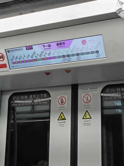 这是一张昆明地铁3号线车厢内部的实拍图，上方电子显示屏清晰展示线路信息：当前为3号线，下一站是梁家河站，同时列出了该线路从西山公园到东部汽车站的完整站点列表，并标注“对侧开门”的乘车提示；车门区域贴有多组中英双语安全指引标识，包括“灯闪/铃响请勿上下车”“小心站台间隙”“禁止倚靠”等，还有“使用电子设备禁止外放声音”的规则提示，整体是公共交通内的标准化乘车指引场景，无谐音梗或双关内容。
