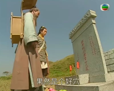 这是一张古装剧的户外场景截图，画面里有两位古装打扮的男子，一位背着风水勘测器具，两人站在一座刻有红字的墓碑前，墓碑前摆放着水果祭品，下方字幕显示“果然是个好穴”，结合场景可知是风水先生在评价墓穴的风水，此处“好穴”指风水优良的墓穴，无谐音或双关含义。背景是晴朗蓝天与草坡，整体呈现出古装剧中勘测墓穴风水的严肃场景。