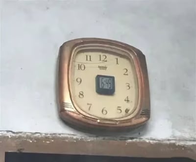 图片中展示了一款独特的挂钟，整体呈现复古风格：铜色金属边框略带磨损，奶油色表盘上印有黑色阿拉伯数字1至12，保留了传统机械钟的经典设计元素。然而，表盘正中央被嵌入了一块小型电子显示屏，清晰显示着数字时间“6:49”，形成传统指针式时钟外观与现代数字计时功能的鲜明混搭。这种改造直观体现了“传统工艺与现代科技的无机结合”主题，既有老式钟表的质感，又具备电子钟的精准读时特性。