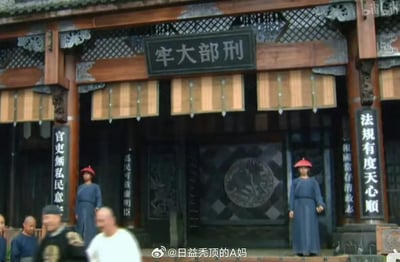 这是一张清代题材影视剧的横版截图，画面呈现了名为“刑部大牢”的清代官署建筑，正门上方悬挂“刑部大牢”牌匾，两侧立柱与墙体上挂有“法规有度天心順”“官吏無私民意安”“為民可效廉明臣”等彰显吏治理念的对联。门口有两名头戴红顶官帽、身着蓝色官服的差役严肃站立值守，前景处有几位清代装扮的官员、百姓正从门前走过，整体场景还原了古代官署的威严氛围，无谐音梗或双关内容。