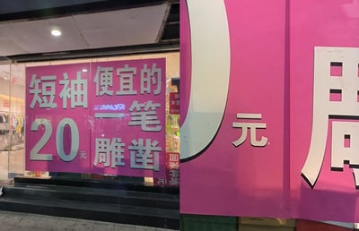 图片展示了一家服装店的店面，左侧玻璃橱窗上贴着大幅粉色促销海报，上面用白色大字写着“短袖 20元 便宜的 一笔 雕凿”。右侧是店铺外墙的一部分，同样为粉色背景，上面有巨大的白色“0元”字样和部分“周”字。海报中的“一笔雕凿”可能是商家故意使用的谐音或错别字来吸引顾客注意，实际想表达“便宜得很”或类似含义。店内可以隐约看到挂着的服装。