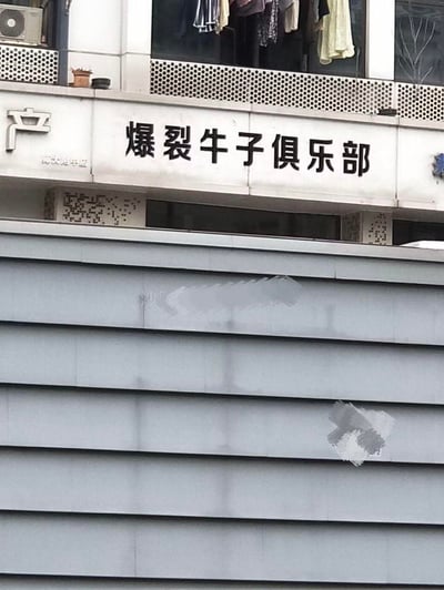 这是一张线下店铺招牌的实拍照片，招牌安装在多层建筑的外墙上，主体文字为「爆裂牛子俱乐部」，左侧还有部分「产」字标识以及「师大附中店」的小字。建筑上方的阳台区域晾晒着多件衣物，招牌下方的墙面有两处被涂抹遮挡的痕迹。该店铺名称使用了网络语境中的低俗谐音梗「牛子」，指代男性生殖器官，用在店铺名称里显得十分奇葩、猎奇，容易引发网友关注与讨论。