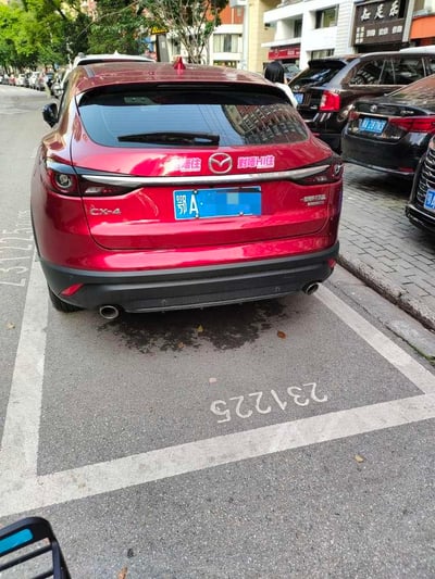 图片展示了一辆红色马自达CX-4汽车的车尾视角，车辆停放在城市街道的划定停车位内。车尾牌照为蓝色底的“鄂A”（湖北武汉）牌照，牌照部分区域被模糊处理。后挡风玻璃上贴有白色文字贴纸，左侧为“喝住”，右侧为“封喝+住”。停车位地面用白色涂料标有“23V225”编号。周围环境可见多辆停放的其他车辆、路边树木及临街商铺建筑，呈现典型的城市居民区或商业区街景。