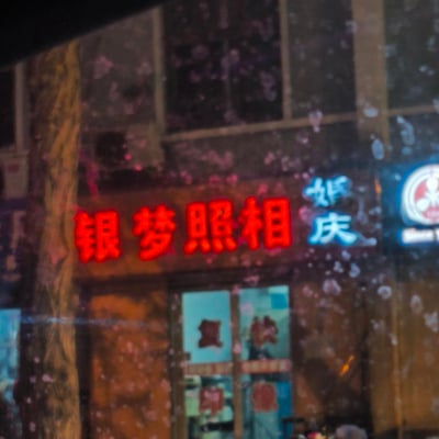 夜晚拍摄的一家名为"银梦照相"的店铺招牌，红色的霓虹灯字样在夜色中格外醒目。旁边还有蓝色的"婚庆"字样，表明这家店可能提供婚纱摄影服务。照片似乎是透过带有雨滴的窗户拍摄的，画面上有许多圆形的光斑，营造出朦胧的氛围。店铺的玻璃门上还能看到一些模糊的红色文字。