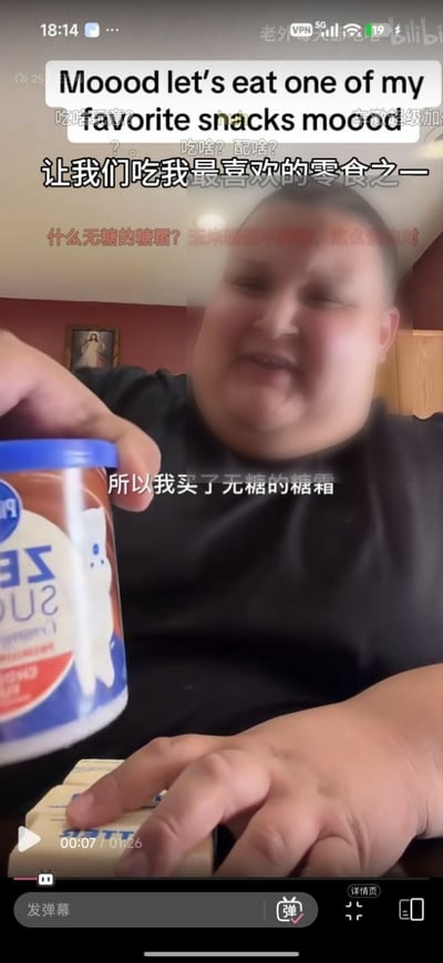 这是一张B站的竖屏视频截图，画面中是一名体型较胖的国外男性博主，正笑着举着一罐印有ZERO SUGAR字样的无糖糖霜产品向镜头展示。画面上方有英文配文，下方配有对应中文字幕，字幕说明博主表示要吃自己喜欢的零食，所以买了无糖的糖霜。画面底部保留了B站的播放进度条、发弹幕控件等界面元素，还能看到部分残留的弹幕内容，核心笑点在于“无糖”和“糖霜”本身属性的矛盾，调侃了部分所谓无糖健康食品的噱头属性。