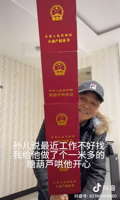 这是一张抖音视频截图，画面中一位戴着灰色带标的帽子、穿着带毛领外套的老人，开心地举着一串用多本中华人民共和国不动产权证书、房屋所有权证串成的“糖葫芦”，配文写道“孙儿说最近工作不好找，我给他做了个一米多的糖葫芦哄他开心”。用将贵重房产证书做成糖葫芦的创意方式，以幽默又硬核的反差感，表达长辈对面临职场压力晚辈的安慰与疼爱，氛围暖心又搞笑。
