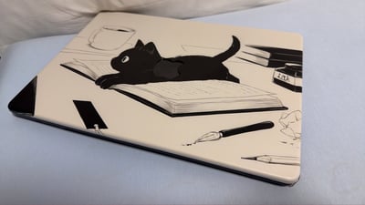 这是一张实拍的MacBook笔记本电脑保护壳展示图，保护壳底色为米白色，采用黑白手绘插画风格：画面中心是一只呆萌的黑色小猫趴在摊开的书本上，设计师巧妙将MacBook本身的苹果logo与黑猫的身体部位融合，毫无违和感；画面周围还搭配绘制了咖啡杯、堆叠的书籍、标有“ink”字样的墨水瓶、蘸水笔、铅笔等文具元素，整体设计可爱充满巧思，保护壳被放置在浅蓝色的平整表面上进行拍摄。