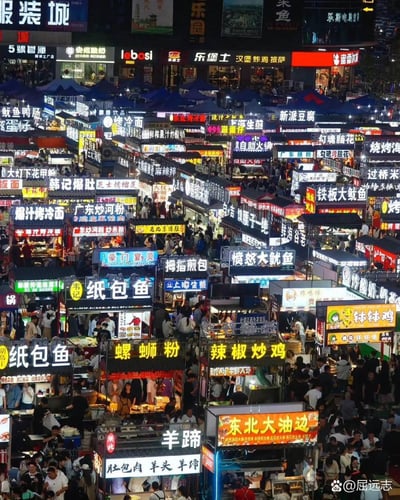 这是一张夜晚的大型夜市美食街实拍图，密密麻麻的小吃摊位鳞次栉比，各色发光的招牌醒目亮眼，涵盖了纸包鱼、螺蛳粉、烤冷面、爆肚、铁板鱿鱼、钵钵鸡等来自全国各地的特色美食，摊位间人头攒动，挤满了前来觅食的人群，整个场景充满浓郁的市井烟火气，生动展现出夜市热闹喧嚣的繁华氛围。