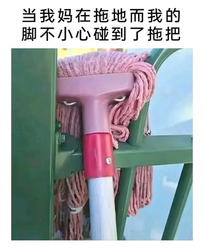 这是一张趣味生活梗图，采用顶配底文的图文混排形式，顶部配文为“当我妈在拖地而我的脚不小心碰到了拖把”，下方主体是一张实拍的粉色拖把的照片，拖把头部的金属卡扣与散开的粉色拖布组合在一起，视觉上形成了瞪着眼睛、满脸怒气的人脸效果，用拟人化的拖把生动指代被打扰拖地的妈妈的神态，精准还原了很多人童年时不小心打断家长做家务时被恶狠狠瞪视的经典场景，很有共鸣感。