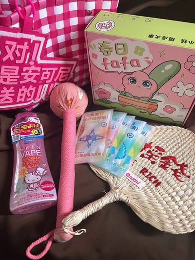 一张展示春日主题礼品套装的照片，包含多个可爱风格的物品。画面中央是粉色和绿色相间的「春日fafa」礼盒，盒上印有卡通盆栽形象，周围摆放着粉色格子纹手提篮、印有「对! 是安可尼送的」字样的粉色布制标语牌、Hello Kitty图案的驱蚊液、粉色布艺玩具锤、几片冰凉贴以及一把带有红色文字的草编扇子。整体色调以粉色为主，充满春日气息和少女心。